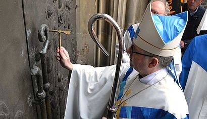 Bischof Kohlgraf steht vor dem geschlossenen Pilgerportal. Er schlägt mit einem goldenen Hammer in der rechten Hand gegen das Portal, in der linken Hand hält er seinen Bischofsstab.