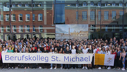 Eine große Gruppe Schüler steht vor dem Berufskolleg. Die erste Reihe hält ein Transparent mit dem Namen der Schule, andere Schüler halten die Flagge Italiens und des Vatikanstaats.