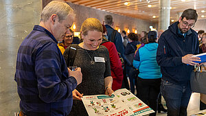 Ein Mann und eine Frau stehen nebeneinander und schauen gemeinsam in eine Zeitschrift. Im Hintergrund stehen viele Menschen. Ein Mann und eine Frau stehen nebeneinander und schauen gemeinsam in eine Zeitschrift. Im Hintergrund stehen viele Menschen.