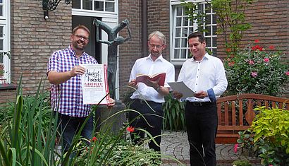 Drei Männer stehen im Innenhof des Priesterhauses. Bastian Rütten hält ein Plakat, Romano Giefer und Fabian Matussek blicken in aufgeschlagene Bücher in ihren Händen.