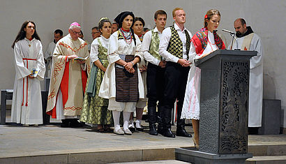 Eine Frau in traditioneller kroatischer Kleidung steht am Ambo der Repelener Kirche, hinter ihr stehen weitere traditionell gekleidete Menschen, um die Fürbitten vorzulesen. Neben einer Messdienerin stehend betet im Hintergrund Weihbischof em. Dieter Geerlings die Fürbitten mit.