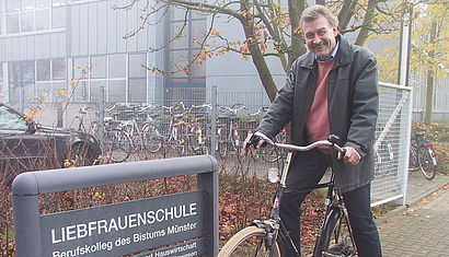 Bernd van Essen sitzt auf einem Fahrrad vor dem Namensschild der Schule.