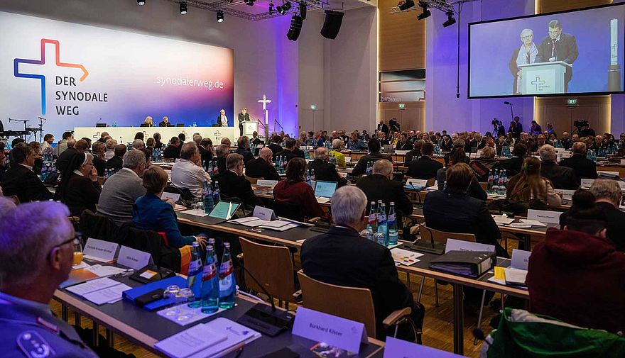 Viele Menschen sitzen an langen Tischen in einem Konferenzsaal.