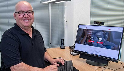 Ein Mann sitzt an einem Computer und lächelt in die Kamera.