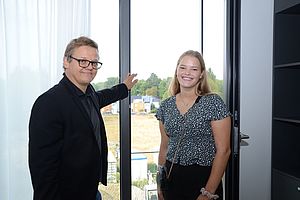 Eine Frau und ein Mann stehen in einem Zimmer an einem geöffneten Fenster. Der Mann weist mit der Hand nach draußen. Eine Frau und ein Mann stehen in einem Zimmer an einem geöffneten Fenster. Der Mann weist mit der Hand nach draußen.