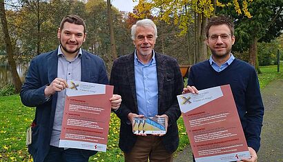 Drei Männer stehen im Freien, zwei halten ein Plakat, einer eine Postkarte.