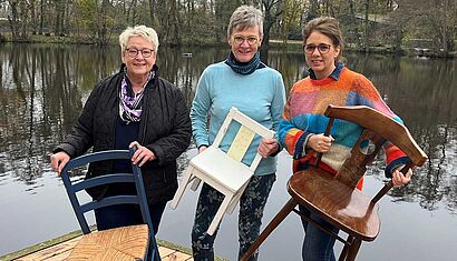 Drei Frauen stehen vor einem Gewässer, sie halten jeweils einen Stuhl in den Händen.