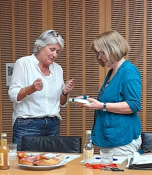 Zwei Frauen stehen an einem Tisch in einem Konferenzraum. Die Frau links trägt eine weiße Bluse und Jeans und gestikuliert im Gespräch. Die Frau rechts trägt eine türkisfarbene Bluse und hält ein Geschenk in den Händen. Zwei Frauen stehen an einem Tisch in einem Konferenzraum. Die Frau links trägt eine weiße Bluse und Jeans und gestikuliert im Gespräch. Die Frau rechts trägt eine türkisfarbene Bluse und hält ein Geschenk in den Händen.
