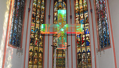 Das Kreuz des Künstlers Ludger Hinse hängt vor den bunten Kirchenfenstern im Altarraum der Marienkirche in Recklinghausen, es leuchtet in allen Regenbogenfarben.