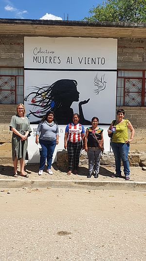 Fünf Frauen stehen auf einer mexikanischen Straße vor einem an einer Hauswand hängenden, großen Plakat. Darauf abgebildet sind eine Frau und eine fliegende Taube.