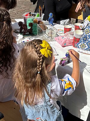 Ein Mädchen mit geflochtenen Haaren und gelber Schleife bemalt an einem Tisch eine kleine Figur. Auf dem Tisch stehen weitere Figuren, Farben und Pinsel.