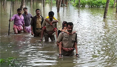 Mehrere Menschen waten durch hüfthohes, braunes Wasser. Zwei Männer tragen jeweils eine weitere Person auf ihren Rücken.