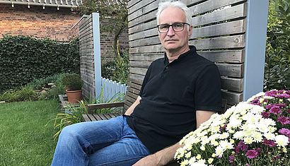 Martin Schmitz sitzt mit Jeans und Pullover bekleidet in einem Garten auf einer Bank