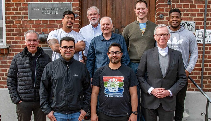 Neun Männer stehen vor einem Gebäude, auf einem Schild an der Mauer steht "Franziskanerkloster Mörmter".