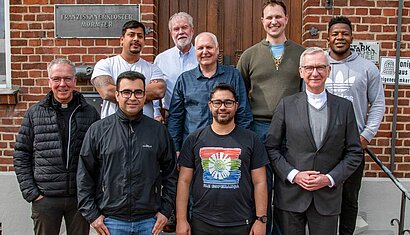 Neun Männer stehen vor einem Gebäude, auf einem Schild an der Mauer steht "Franziskanerkloster Mörmter".