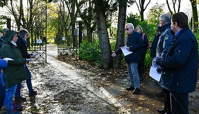 Mehrere Menschen stehen vor einem geöffneten Tor, das auf einen Friedhof führt.