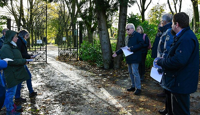 Mehrere Menschen stehen vor einem geöffneten Tor, das auf einen Friedhof führt.