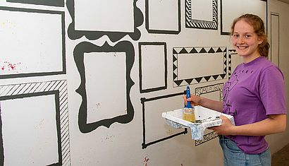 Eine junge Frau hält einen Pinsel und steht an einer Wand mit aufgemalten Bilderrahmen.