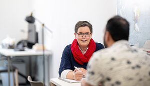 Gudrun Blumenkemper sitzt in ihrem Büro an einem Tisch und spricht mit einem ihr gegenüber sitzenden Mann. In der Hand hält sie ienen Kugelschreiber, auf dem Tisch liegt ein Schreibblock. Gudrun Blumenkemper sitzt in ihrem Büro an einem Tisch und spricht mit einem ihr gegenüber sitzenden Mann. In der Hand hält sie ienen Kugelschreiber, auf dem Tisch liegt ein Schreibblock.