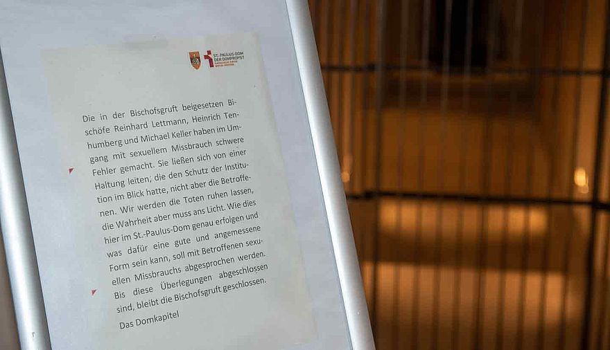 Vor einem verschlossenen Gitter steht eine Stele mit einem erklärenden Text.