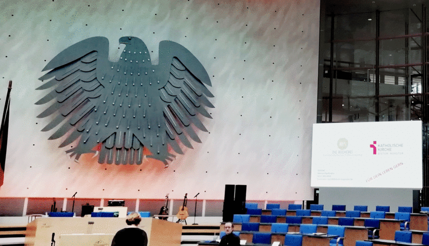 Neben dem Bundesadler im ehemaligen Plenarsaal in Bonn ist auf einer Leinwand das Logo des Bistums zu sehen.