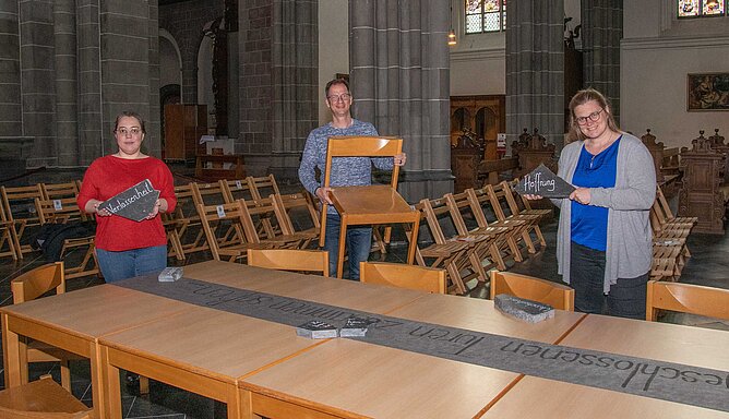 Christiane Flüchter, Matthias Heinrich und Anna Zoll stehen im St. Viktor Dom. Die Frauen halten Steintafeln mit einer Aufschrift, der Mann einen Stuhl in den Händen.