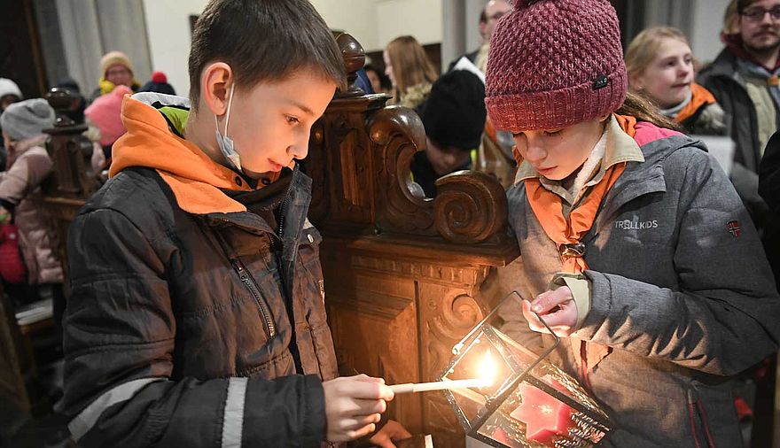 Ein Junge hält eine brennende Kerze in der Hand, mit der er das Windlicht eines Mädchens anzünden möchte.