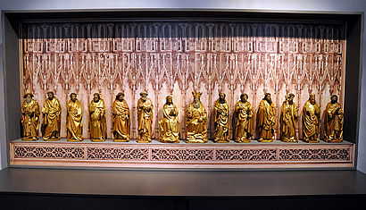 14 goldfarbene Heiligenfiguren aus Holz stehen nebeneinander vor einer Wand, die wie ein mittelalterlicher Altar gestaltet ist, im Museum.