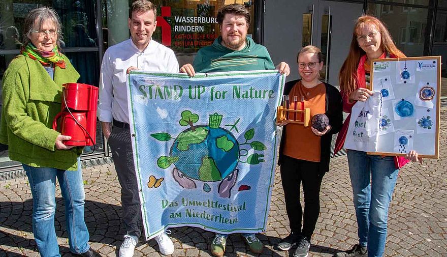 Drei Frauen und zwei Männer halten verschiedene Gegenstände, unter anderem ein Werbebanner für ein Umweltfestival.