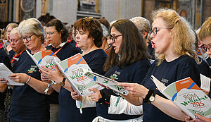 Frauen und Männer singen im Chor mit einheitlichen dunkelblauen Shirts bekleidet und Chorbüchern in der Hand.