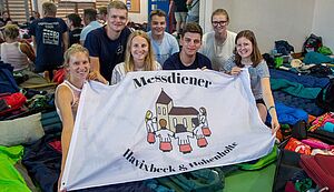 In einem Raum, dessen Boden mit Schlafsäcken und Gepäckstücken bedeckt ist, zeigen Lea Kleine-Vornholt, Lenn Fehmer, Pia Farber, Maximilian Pape, Stefan Mühlenbeck, Sarah Lülf und Johanna Pape eine Fahne mit der Aufschrift "Messdiener Havixbeck & Hohenholte". In einem Raum, dessen Boden mit Schlafsäcken und Gepäckstücken bedeckt ist, zeigen Lea Kleine-Vornholt, Lenn Fehmer, Pia Farber, Maximilian Pape, Stefan Mühlenbeck, Sarah Lülf und Johanna Pape eine Fahne mit der Aufschrift "Messdiener Havixbeck & Hohenholte".