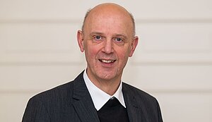 Das Bild zeigt Kurt Schulte. Er trägt eine dunkle, fein gestreifte Jacke, ein weißes Hemd und einen schwarzen Pullover. Der Hintergrund ist hell und schlicht gehalten.