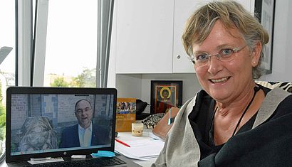 Dr. Elke Kleuren-Schryvers sitzt an einem Schreibtisch und blickt in die Kamera, auf dem Tisch steht ein aufgeklappter Laptop, auf dessen Bildschirm ein Video mit Weihbischof Rolf Lohmann zu sehen ist.