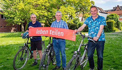 Drei Männer stehen neben ihren Fahrrädern und halten einen roten Schal mit der Aufschrift "leben teilen" fest.