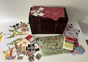 Auf einem weißen Tisch liegt ein geöffnetes Bildungs- und Spielmaterial-Set rund um Kinderrechte. Im Zentrum steht eine dunkelbraune Holztruhe mit Lederriemen. Darauf und daneben sind zahlreiche Materialien ausgebreitet: Karten mit Aufgaben und Symbolen, ein Spielbrett mit Spielfiguren und Würfeln, Bildkarten, Illustrationen von Kindern und Erwachsenen, ein Kartenspiel sowie mehrere Broschüren und Hefte. Die Materialien wirken thematisch geordnet und laden zum gemeinsamen Lernen und Spielen ein. Im Hintergrund befindet sich eine schlichte weiße Wand.