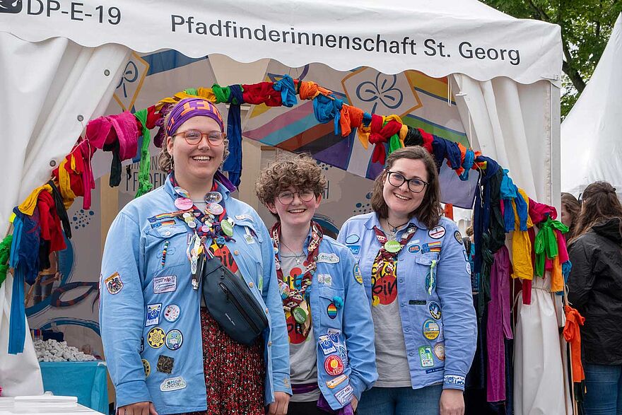 Drei Pfadfinderinnen stehen vor einem Zelt, an dem bunte Tücher hängen.