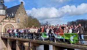 Eine Gruppe von etwa 40 bis 50 Menschen steht nebeneinander auf einer Brücke und blickt in die Kamera. Einige halten mehrere Fahnen und Banner, darunter auch eine Regenbogenflagge. Im Hintergrund steht ein historisches Gebäude mit Turm und Uhr, das an ein Schloss erinnert. Unter der Brücke fließt ein Wassergraben, und darüber ist ein blauer Himmel mit großen Wolken zu sehen.