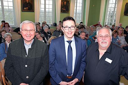 Drei Männer stehen in einem Saal mit dem Gesicht zum Betrachter. Hinter ihnen sitzen mehrere Menschen.