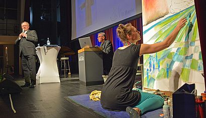 Im Vordergrund mal die Künstlerin Tatjana van Went auf dem Boden der Bühne hockend ein Bild, im Hintergrund steht Wallfahrtsrektor Gregor Kauling.