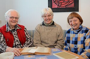 Drei ältere Frauen sitzen gemeinsam an einem Tisch und betrachten ein geöffnetes Buch. Vor ihnen liegen weitere Unterlagen und ein Buch. Im Hintergrund hängt ein Bild mit einem stilisierten Vogel. Drei ältere Frauen sitzen gemeinsam an einem Tisch und betrachten ein geöffnetes Buch. Vor ihnen liegen weitere Unterlagen und ein Buch. Im Hintergrund hängt ein Bild mit einem stilisierten Vogel.