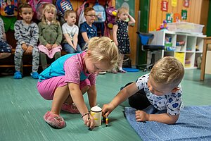 Eine Gruppe von Kindern in einer Kita. Im Vordergrund sitzen ein Mädchen in rosa-pinker Kleidung und ein Junge in weißem T-Shirt mit Sternmuster auf dem Boden. Beide spielen gemeinsam mit kleinen Spielfiguren und einer weißen Kerze. Im Hintergrund sitzen weitere Kinder auf einer Bank und schauen zu, bunte Rucksäcke hängen an der Wand.