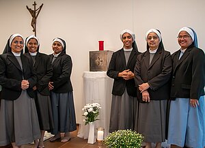 Sechs Ordensschwestern stehen nebeneinander in einem schlicht eingerichteten Raum mit einem Kruzifix an der Wand. Alle tragen graue Kleider mit schwarzem Blazer und Schleier. In der Mitte des Raumes steht ein kleiner Altar mit einem Tabernakel, darüber eine rote Lampe. Auf dem Boden stehen weiße Blumen und eine Kerze. Sechs Ordensschwestern stehen nebeneinander in einem schlicht eingerichteten Raum mit einem Kruzifix an der Wand. Alle tragen graue Kleider mit schwarzem Blazer und Schleier. In der Mitte des Raumes steht ein kleiner Altar mit einem Tabernakel, darüber eine rote Lampe. Auf dem Boden stehen weiße Blumen und eine Kerze.
