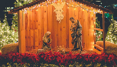 Eine hölzerne Krippe mit einem Engel, Maria, Josef und dem Jesuskind auf einem mit Blumen und Lichterketten geschmückten Platz. 