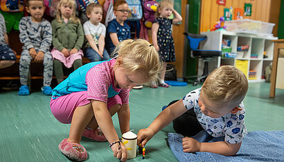 Zwei Kinder spielen in einem Kindergartenraum auf dem Boden mit einer Kerze und einer Figur. Im Hintergrund sind weitere Kinder zu sehen. 