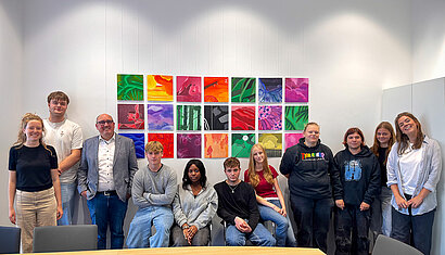 Eine Schulklasse steht/sitzt vor Bildern, die sie selbst gestaltet haben, die hinter ihnen an der Wand hängen.