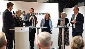 Sechs Personen stehen auf einer Bühne bei einer Podiumsdiskussion im Landtag Nordrhein-Westfalen. Fünf von ihnen stehen hinter zwei Stehtischen mit Mikrofonen und Wassergläsern. Eine Person in der Mitte spricht in ein Mikrofon. Im Hintergrund sind das Logo und die Schrift „Der Landtag Nordrhein-Westfalen“ zu sehen. Das Publikum sitzt im Vordergrund und hört zu.