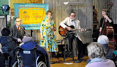 Vor Senioren, die zum Teil im Rollstuhl sitzen, stehen und sitzen vier Musiker, gekleidet im Stil der 1950er-Jahre.