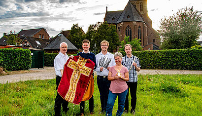 5 Personen stehen auf einer Wiese vor einer Kirche und halten verschiedene Gegenstände wie z.B. ein Messgewand in der Hand.