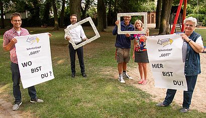 Fünf Personen stehen in einem Park. Zwei halten Plakate fest, drei mit Alufolie umwickelte Fensterrahmen.