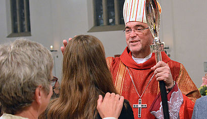 Weihbischof Lohmann spendet einem Mädchen, das von hinten zu sehen ist, das Sakrament der Firmung, indem er die Stirn der Jugendlichen mit Chrisam salbt.